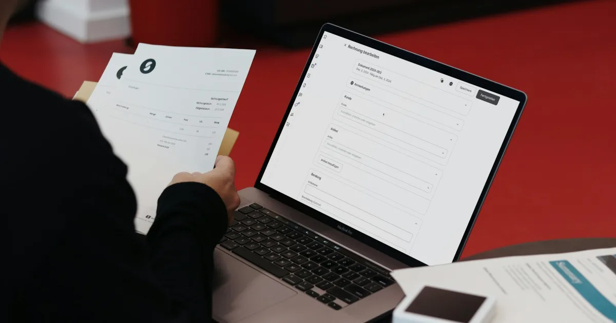 Someone holding paper documents beside an invoicing system open on a screen